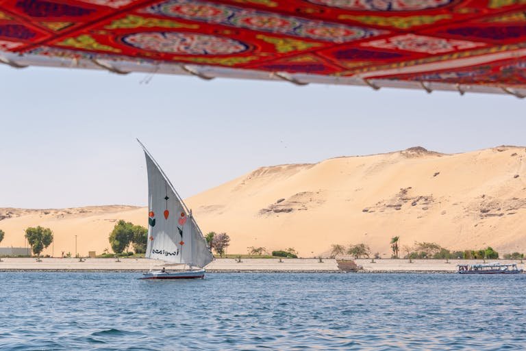 A traditional sailboat sails on the Nile with sand dunes in the background, under a vibrant canopy.