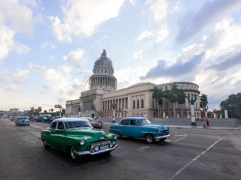Classic cars drive past El Capitolio in Havana, showcasing vintage Cuban architecture and urban life.