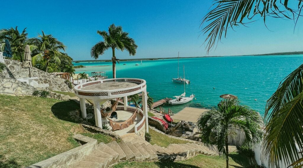 Discover the tranquil paradise of Bacalar Lagoon with palm trees, boats, and a gazebo in stunning blue waters.